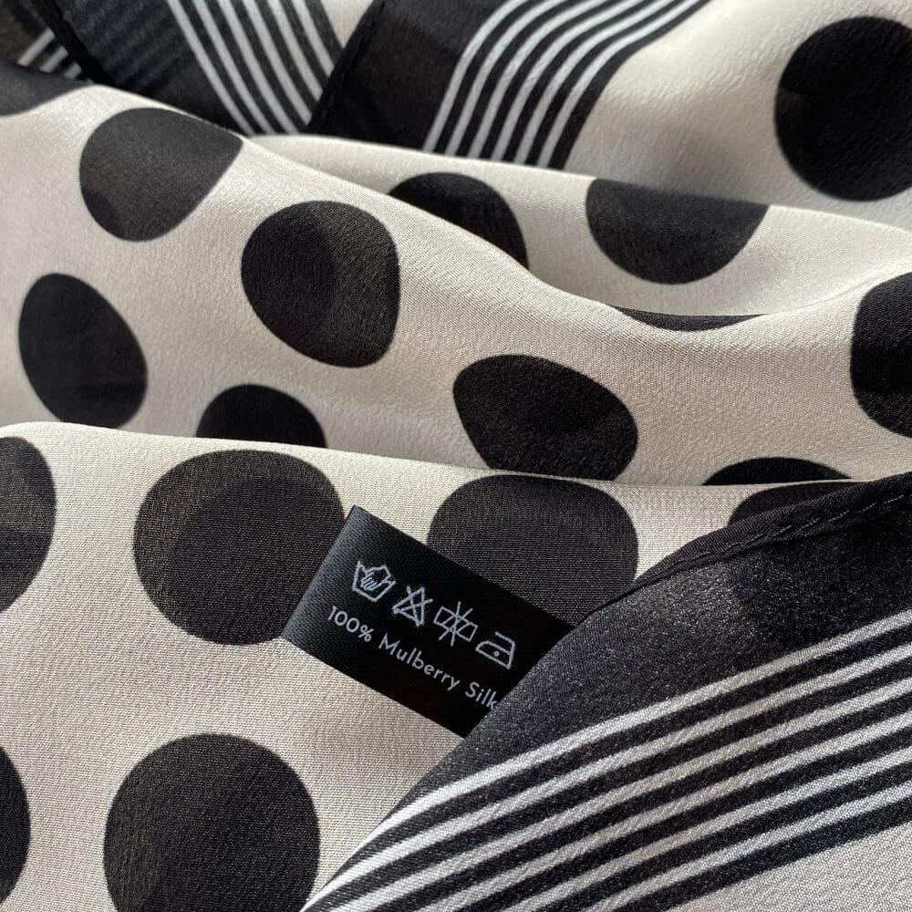 Detail shot of the black and white polka dot Mulberry silk scarf with care label, highlighting the smooth Crepe de Chine fabric and print.