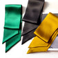 Three mulberry silk satin skinny neck scarf in green, black, and mustard yellow, folded and arranged on a white background with a magazine partially visible.