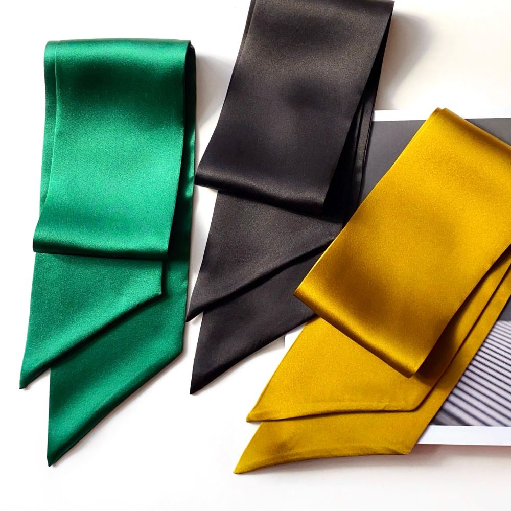 Three mulberry silk satin skinny neck scarf in green, black, and mustard yellow, folded and arranged on a white background with a magazine partially visible.