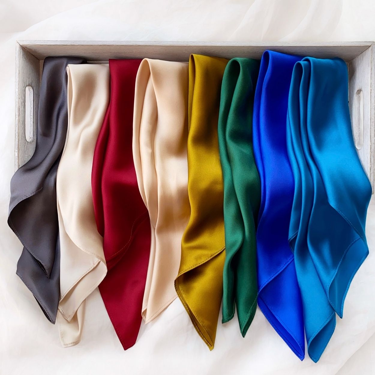 multi colours mulberry silk square scarves arranged side-by-side inside a grey wooden tray. The colours are dark grey, ivory cream, berry red, champagne beige, gold olive, emerald green, royal blue and kingfisher blue.