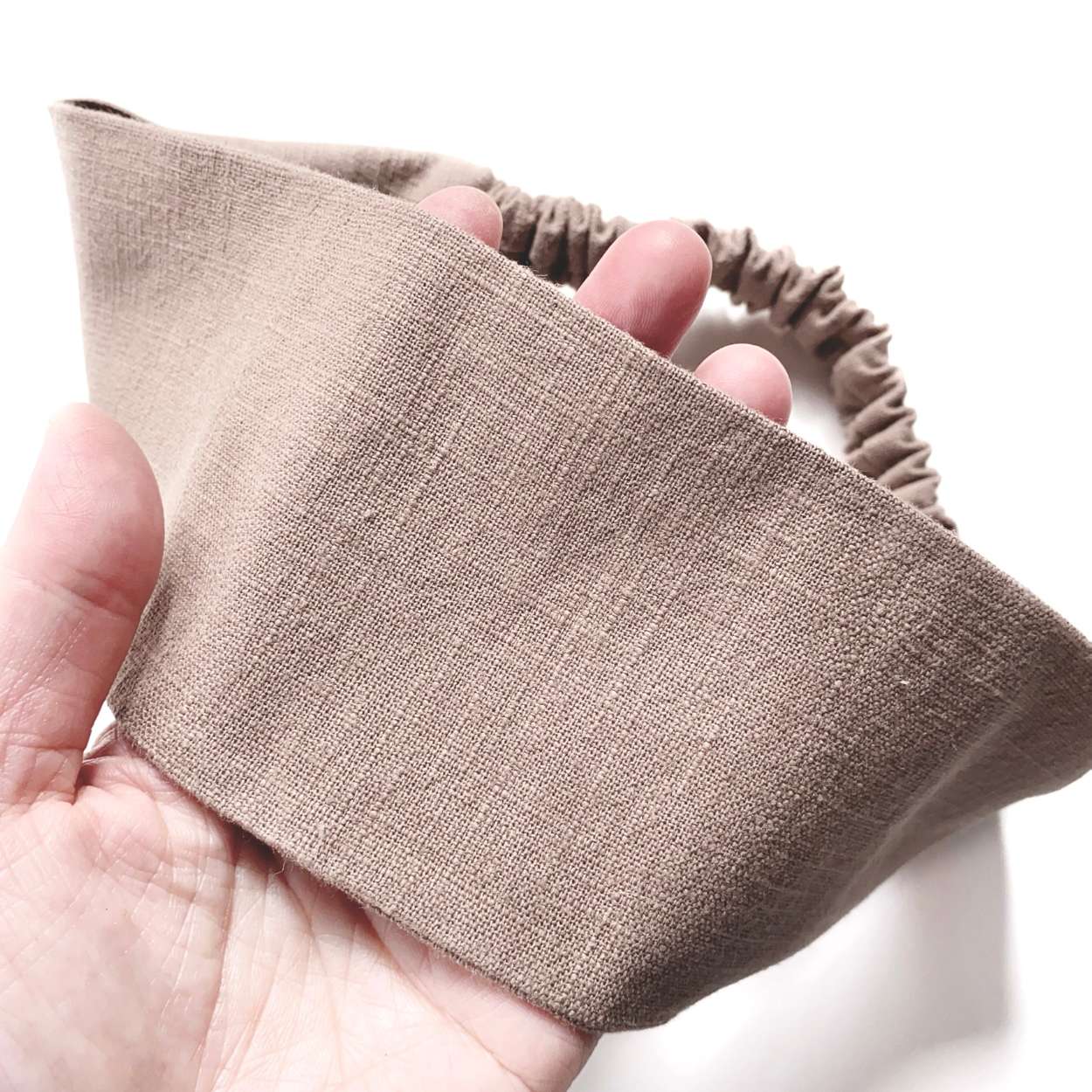 Brown linen fabric headband held in a hand against a white background