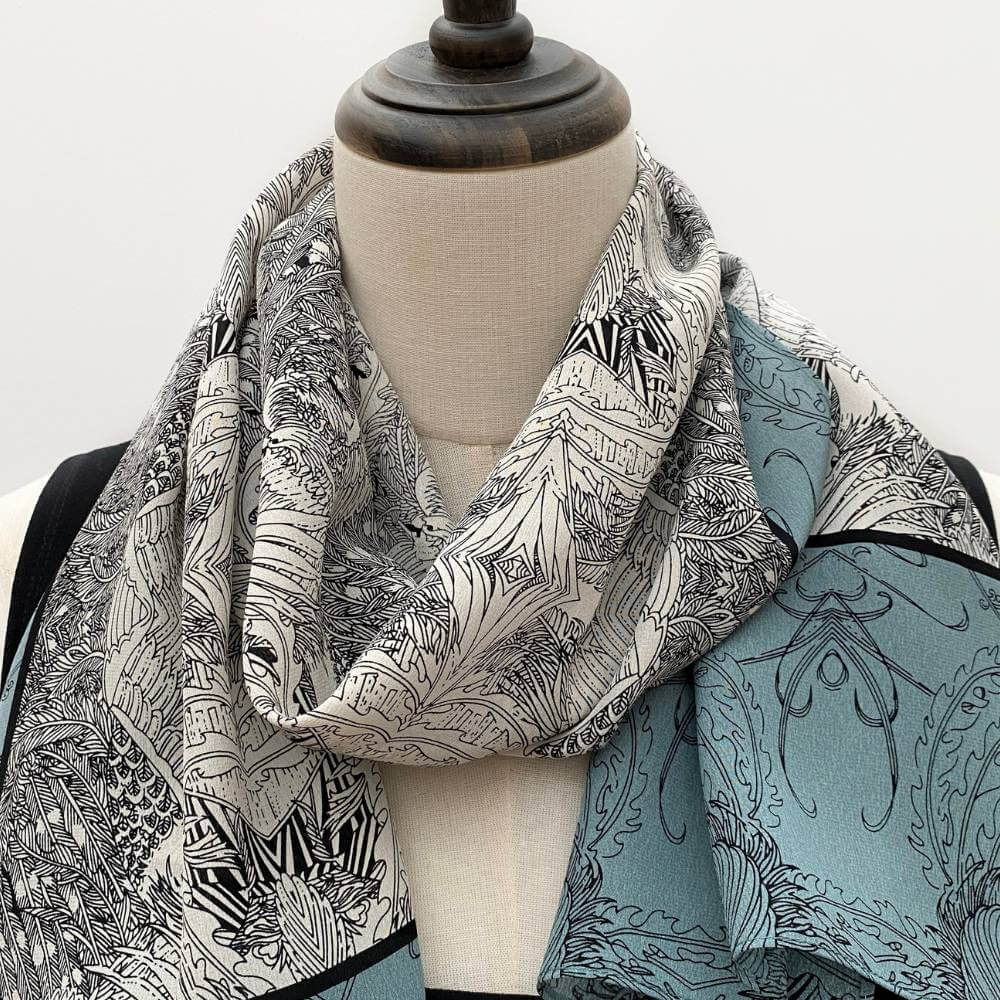 Teal, black and ivory illustrated long silk scarf styled on a mannequin over a black top.