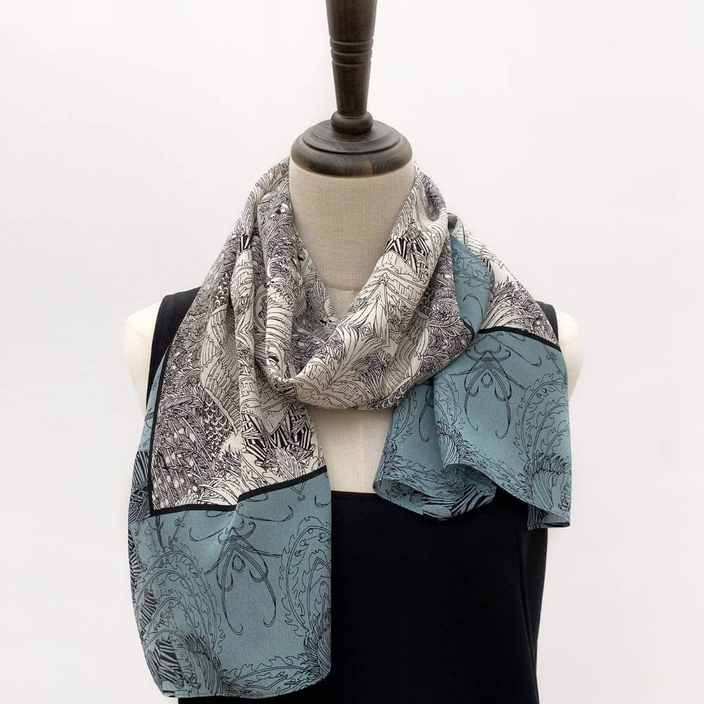 Teal, black and ivory illustrated long silk scarf styled on a mannequin over a black top.