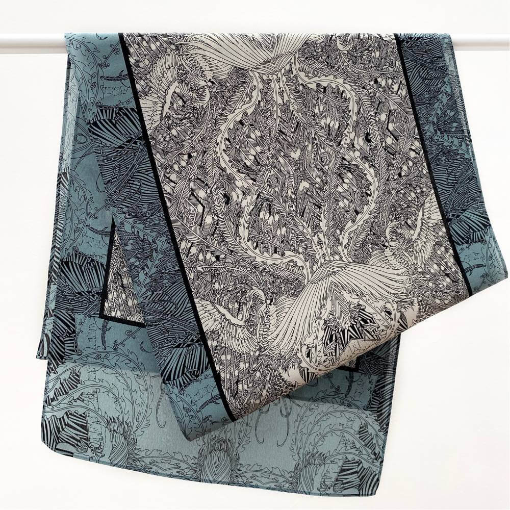 Teal, black and ivory illustrated long silk scarf in crepe de chine, draped over a white hanging bar.
