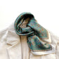 Close-up of a teal green equestrian patterned mulberry silk square neckerchief with gold and silver accents draped over a white jacket on a white background
