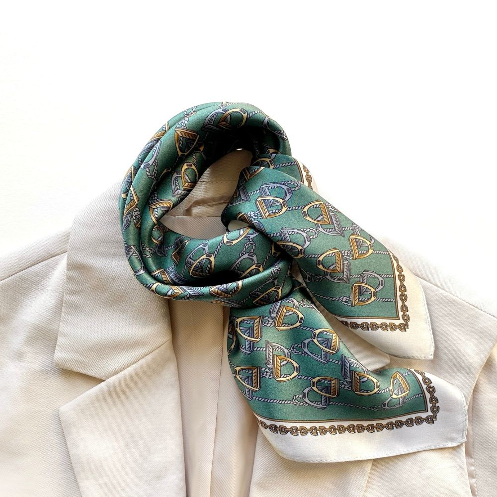 Close-up of a teal green equestrian patterned mulberry silk square neckerchief with gold and silver accents draped over a white jacket on a white background