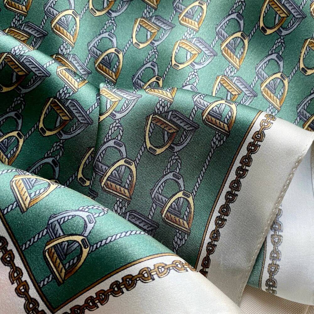 Close-up of a teal green equestrian stirrup patterned mulberry silk square scarf with gold and silver accents