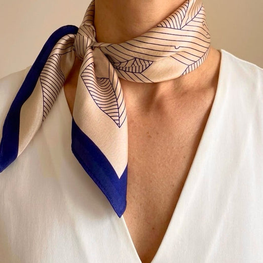Close-up of a dusty rose pink silk scarf with navy blue geometric line art patterns and solid navy borders, tied around a model's neck over a white V-neck top.