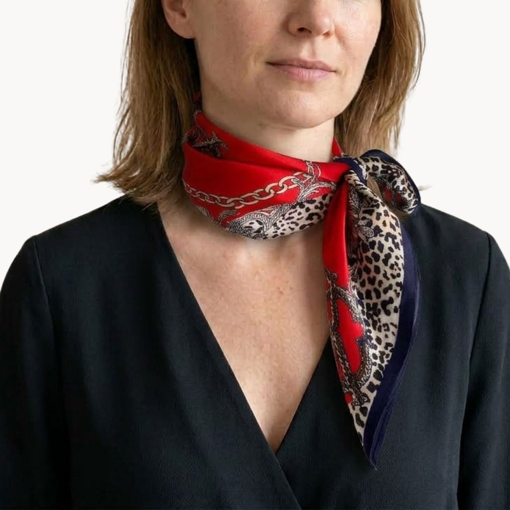A close-up of a woman wearing a red mulberry silk square scarf with a leopard print and chain pattern tied around her neck, paired with a black top against a plain white background.