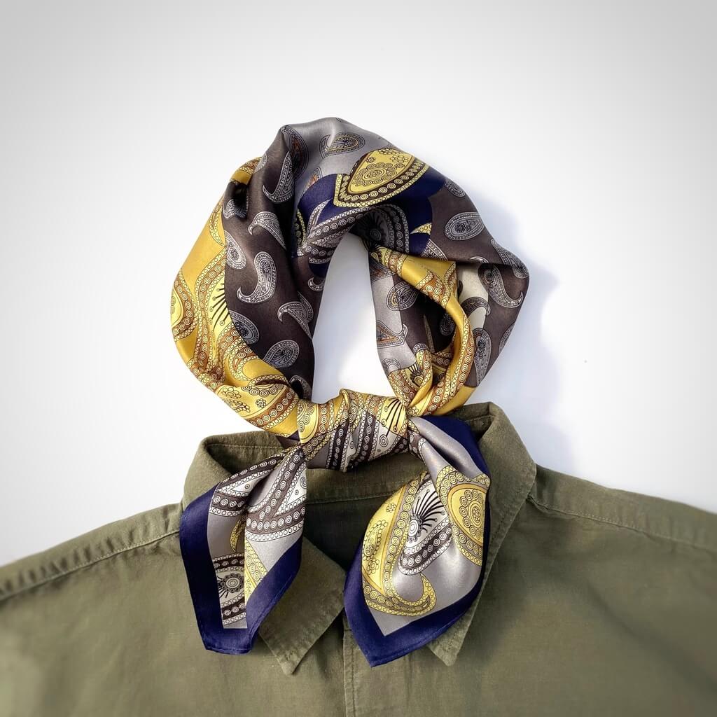 Patterned yellow grey mulberry silk neck scarf draped over a green shirt on a white background